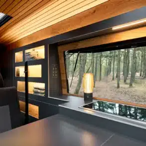 Großes Aufstellfenster im Wohnbereich eines Camper XL mit Blick auf den Wald, moderne Einrichtung mit Holzdecke, Regalen und Beleuchtung