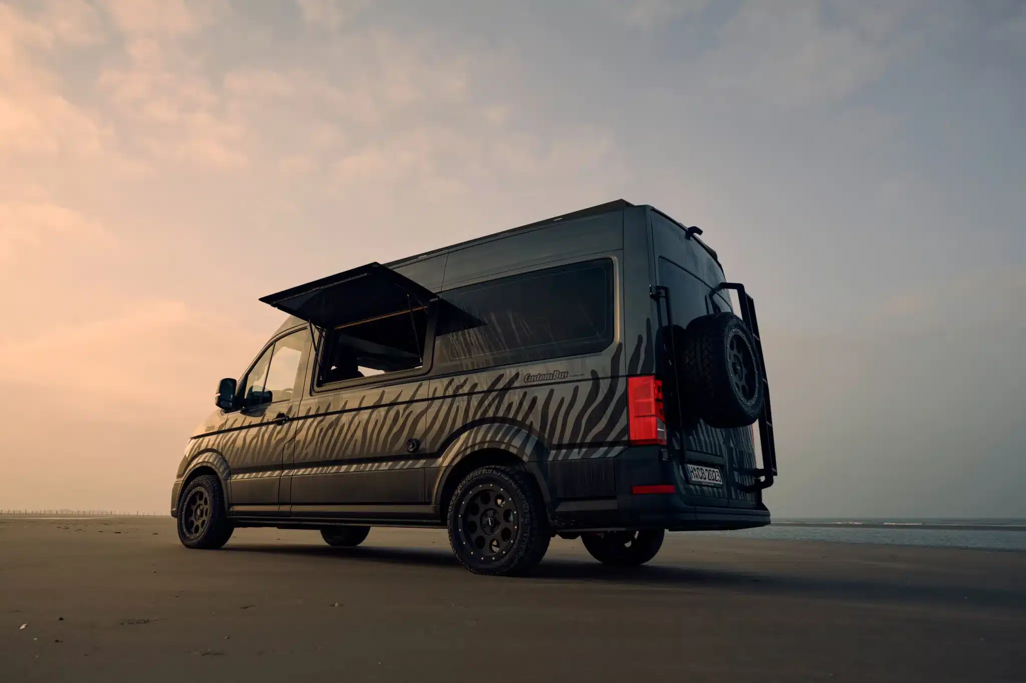 Custom-Bus Camper XL seitlich, am Strand geparkt, mit geöffnetem Aufstellfenster und Heckträger im Sonnenuntergang.