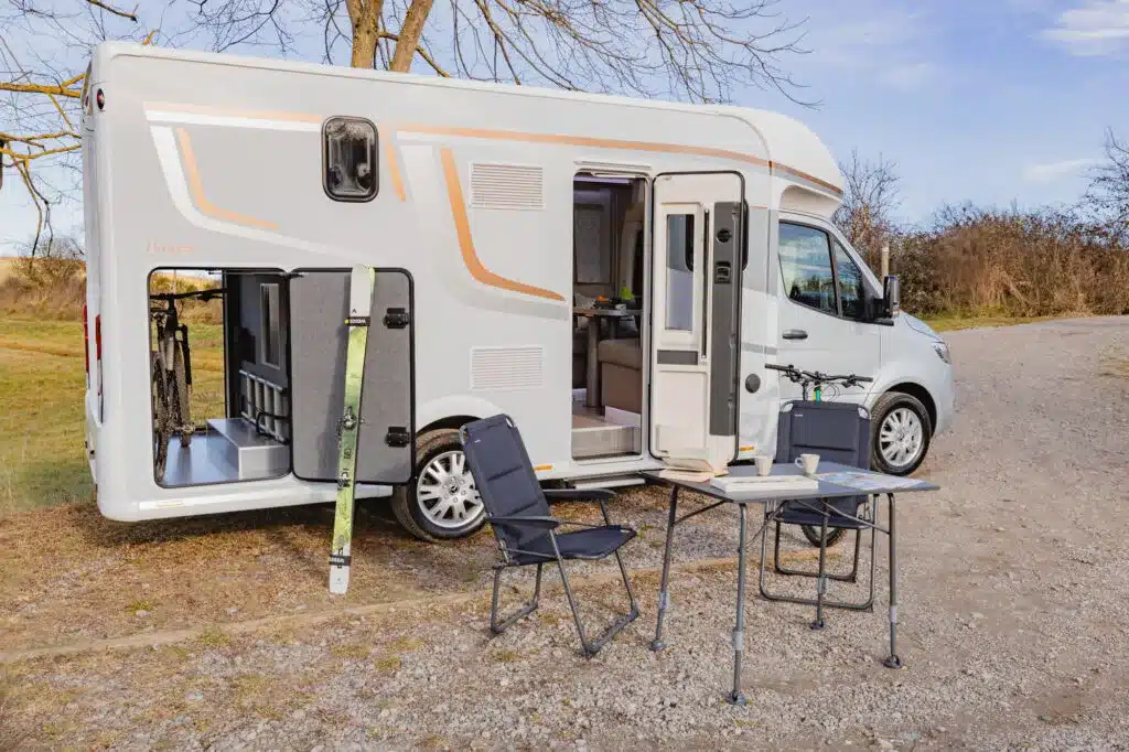 Notin Exclusiv Bruges teilintegrierter Luxus-Camper von außen, mit geöffneter Heckgarage, Klapptisch und Stühlen