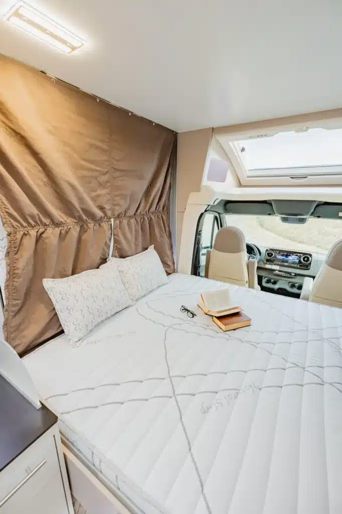Innenansicht des Notin Exclusiv Bruges mit Bett, Kissen, Lesebrille und Buch im Schlafbereich sowie Sicht auf das Panoramadach und das Cockpit.