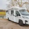 Weißes Notin Vitoria Reisemobil steht auf einem Schotterplatz vor einem See mit ländlicher Umgebung.
