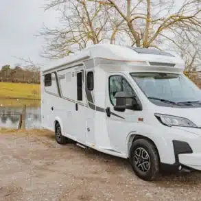 Weißes Notin Vitoria Reisemobil steht auf einem Schotterplatz vor einem See mit ländlicher Umgebung.