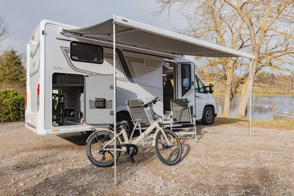 Notin Vitoria Wohnmobil mit aufgeklappter Markise, zwei Campingstühlen und E-Bike auf einem Stellplatz am See
