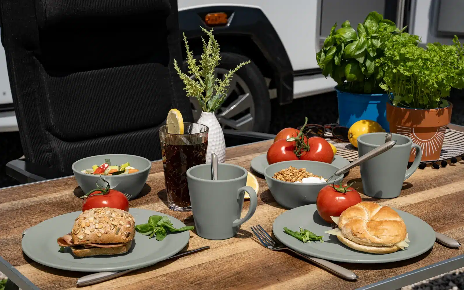Frühstückstisch mit Camping-Geschirr, belegten Brötchen, Tomaten und Pflanzen vor einem Wohnmobil