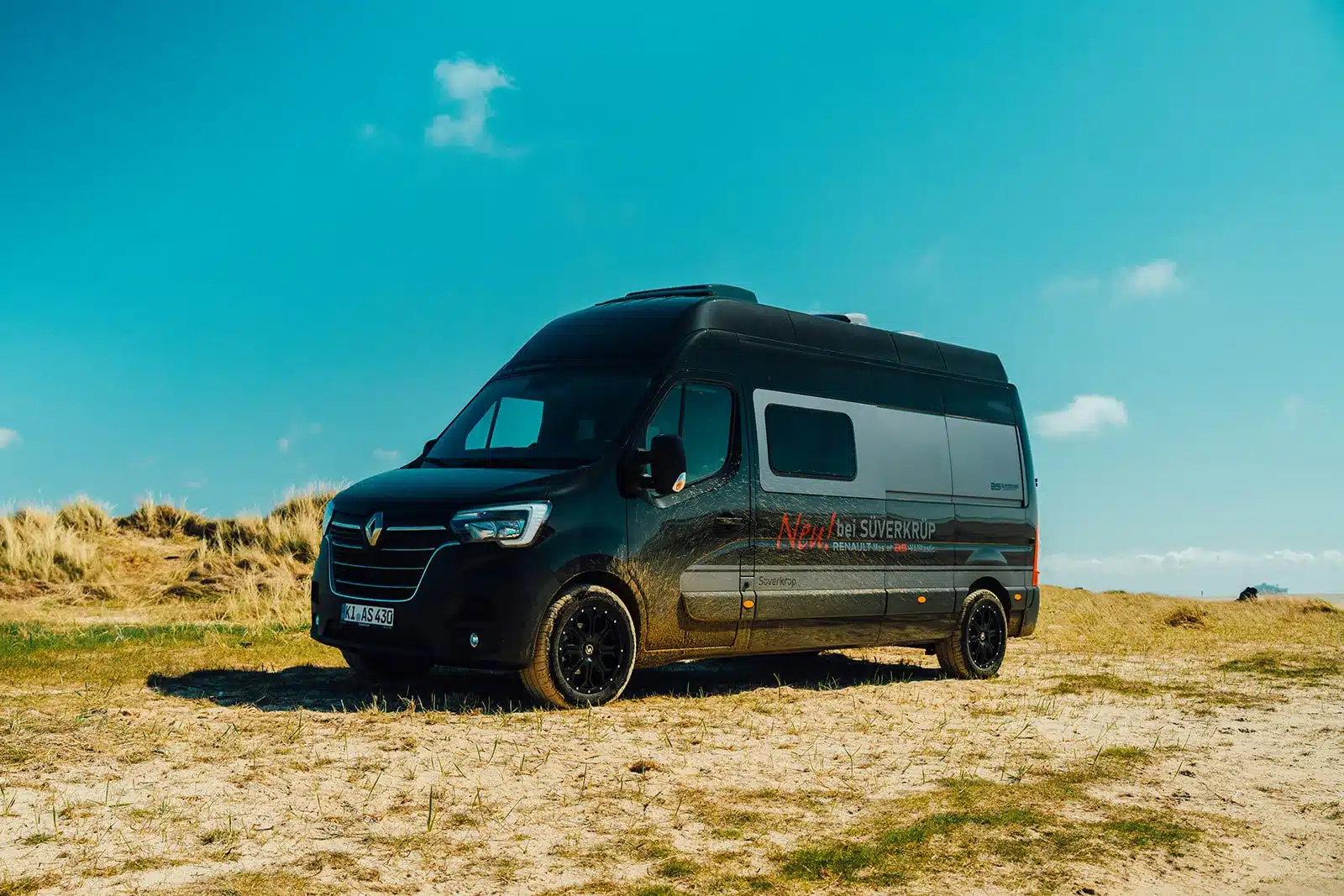 Schwarzer Renault Master VANtastic Kastenwagen steht auf sandigem Untergrund unter blauem Himmel.