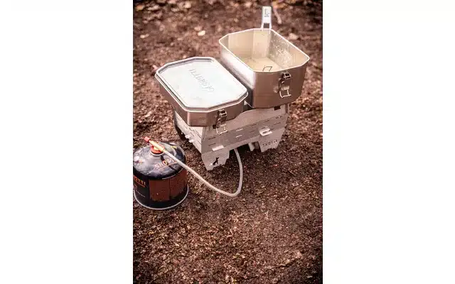 SKOTTI Faltbarer Campinkocher Booster aus Edelstahl mit angeschlossener Gaskartusche und Kochgeschirr im Outdoor-Einsatz auf Waldboden