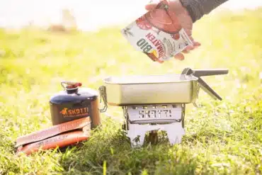 Faltbarer SKOTTI BOOSTER Gaskocher mit Pfanne, Gaskartusche und Kochzubehör beim Kochen im Freien.