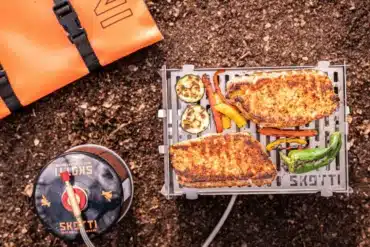 SKOTTI MINI Grill mit gegrilltem Fleisch und Gemüse, angeschlossen an eine Gasflasche; daneben liegt die orange Tragetasche auf Waldboden.