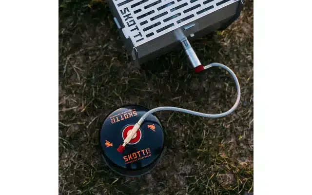 Skotti Schraubkartusche 500 g mit angeschlossenem Gasschlauch an einem Grill auf einer Wiese.