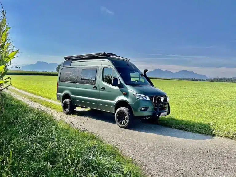 Grüner Mercedes-Benz Sprinter Campervan mit Offroad-Ausstattung auf einem landwirtschaftlichen Weg vor Wiesen und Bergen