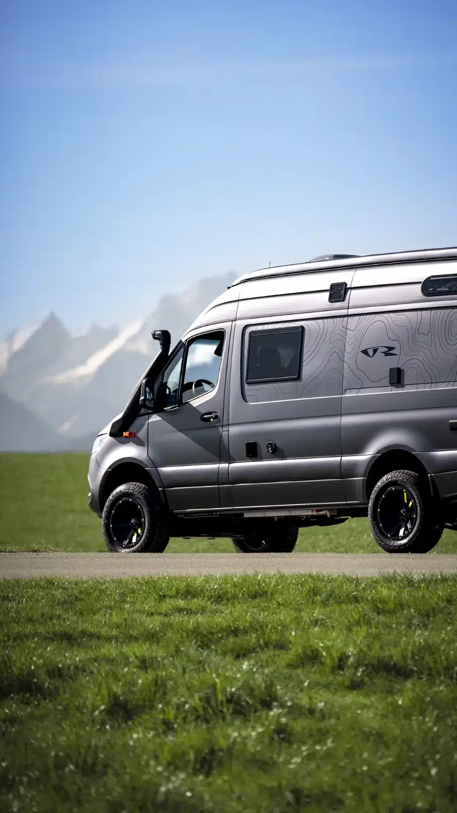 Seitenansicht eines silbergrauen Mercedes-Benz Sprinter 6,0 m Campervans von VR Motorhomes auf einer Wiese mit Alpen im Hintergrund