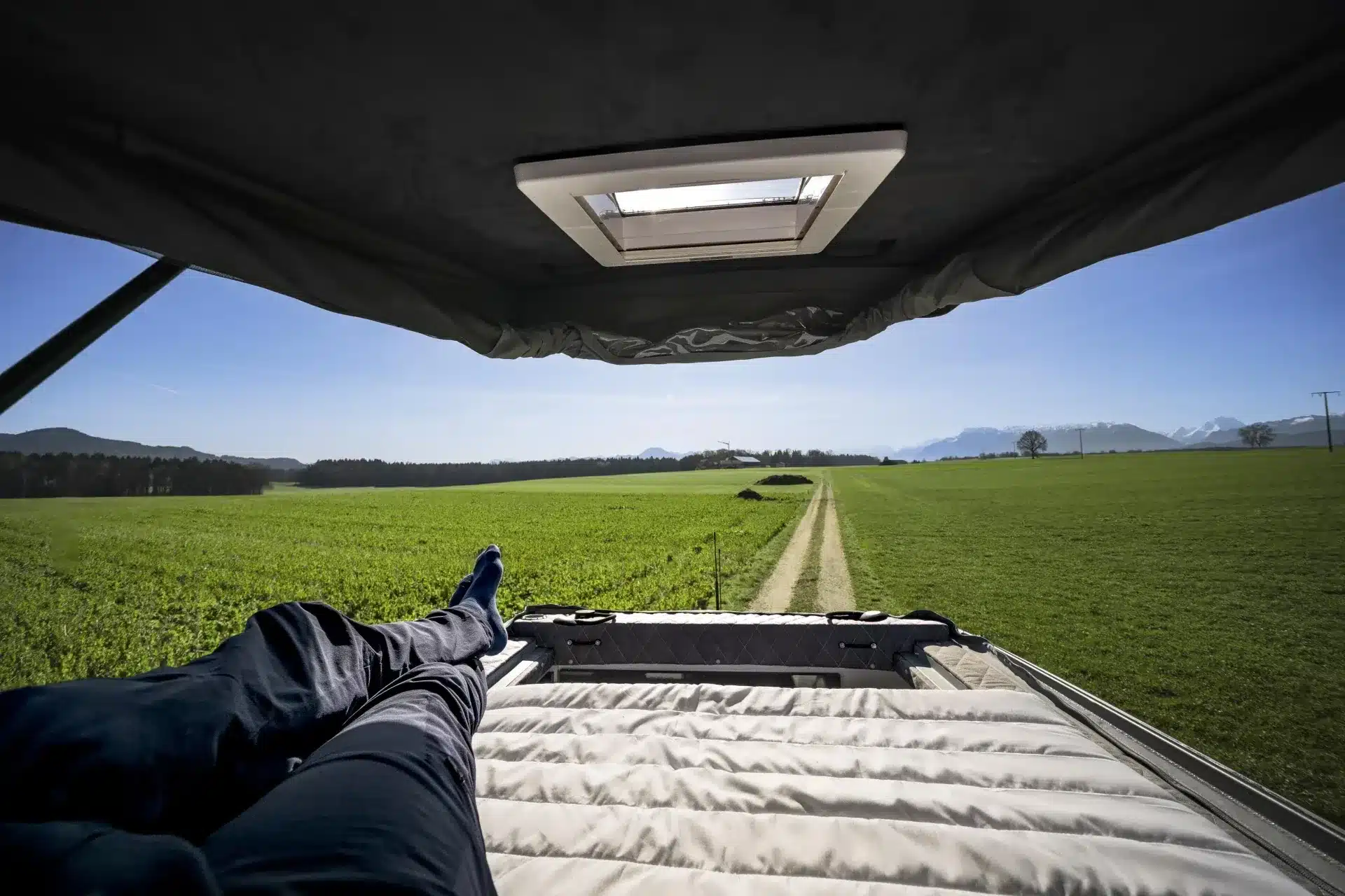 Person liegt entspannt im Aufstelldach eines Kastenwagen-Campers und blickt auf eine grüne Landschaft mit Bergen am Horizont
