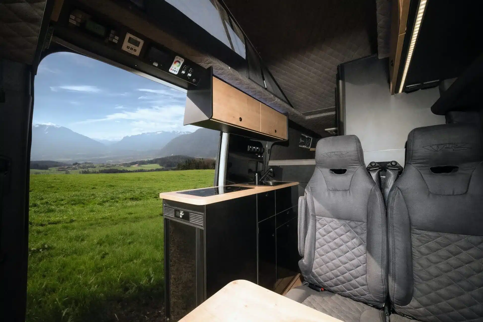 Blick aus dem Sprinter 7 m Camper mit Küchenzeile und Sitzen auf eine weite grüne Wiese und Berge