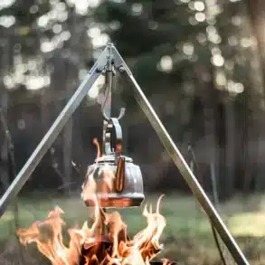 Teekessel aus Edelstahl hängt an Dreibein-Gestell über einem offenen Lagerfeuer im Wald.