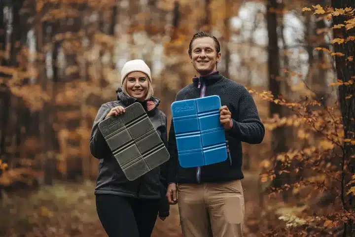 Zwei Personen halten faltbare Thermo-Sitzkissen von Naturbummler, eines in Blau, eines in Grün, im herbstlichen Wald.
