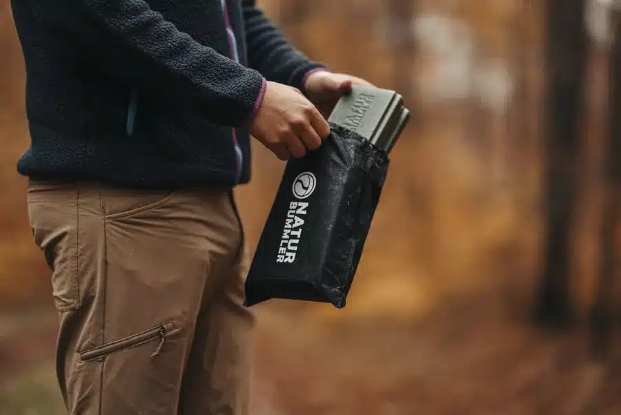 Person verstaut ein faltbares Thermo-Sitzkissen von Naturbummler in eine schwarze Transporttasche im herbstlichen Wald.