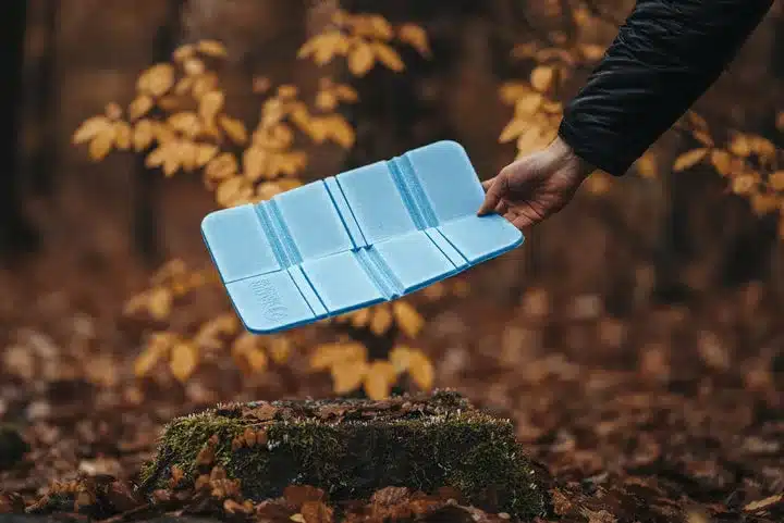 Eine Person hält ein aufgefaltetes, blaues Thermo-Sitzkissen über einen mit Moos bedeckten Baumstumpf im Laubwald.