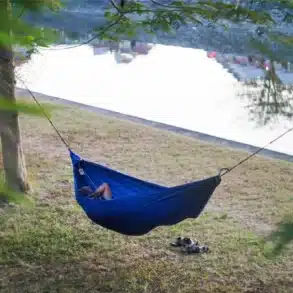 Reisende Person liegt in einer blauen Hängematte zwischen Bäumen an einem Seeufer