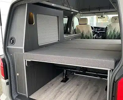 Innenaufnahme eines Transporters mit VA102 Premium Camperausbau, ausklappbarem Bett und dunkler Holzverkleidung.