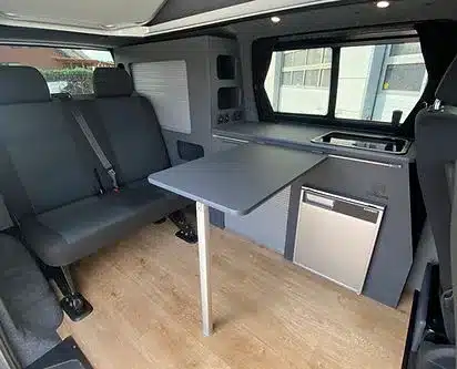Innenansicht eines ausgebauten Transporters mit Sitzbank, Klapptisch, Küchenzeile und Kühlschrank, VA102 Premium Ausbau