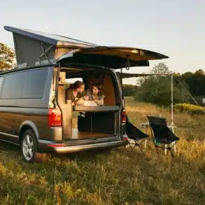 Ein Volkswagen T6.1 Campervan mit aufgeklapptem Dach, geöffneter Heckklappe und zwei Personen im Innenraum auf einem Campingplatz am Wasser