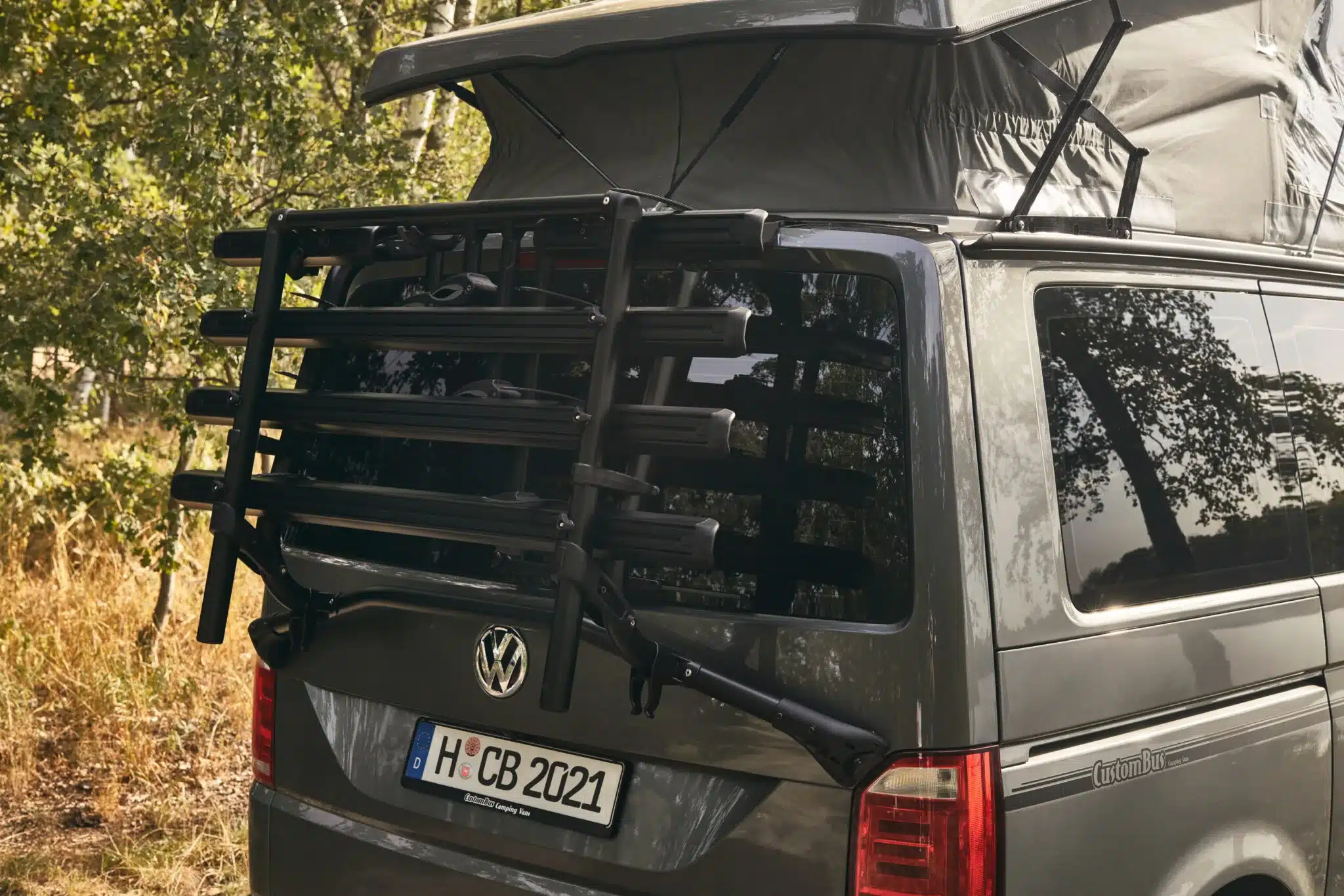 Heck eines VW T6.1 Custom-Bus mit montiertem Fahrradträger und geöffnetem Aufstelldach im Wald