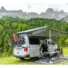 Campingbus mit ausgefahrener Fiamma Markise, Fahrrädern und zwei Frauen am Campingtisch in den Bergen.