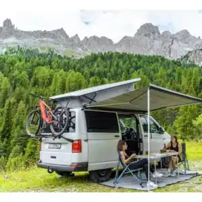 Campingbus mit ausgefahrener Fiamma Markise, Fahrrädern und zwei Frauen am Campingtisch in den Bergen.