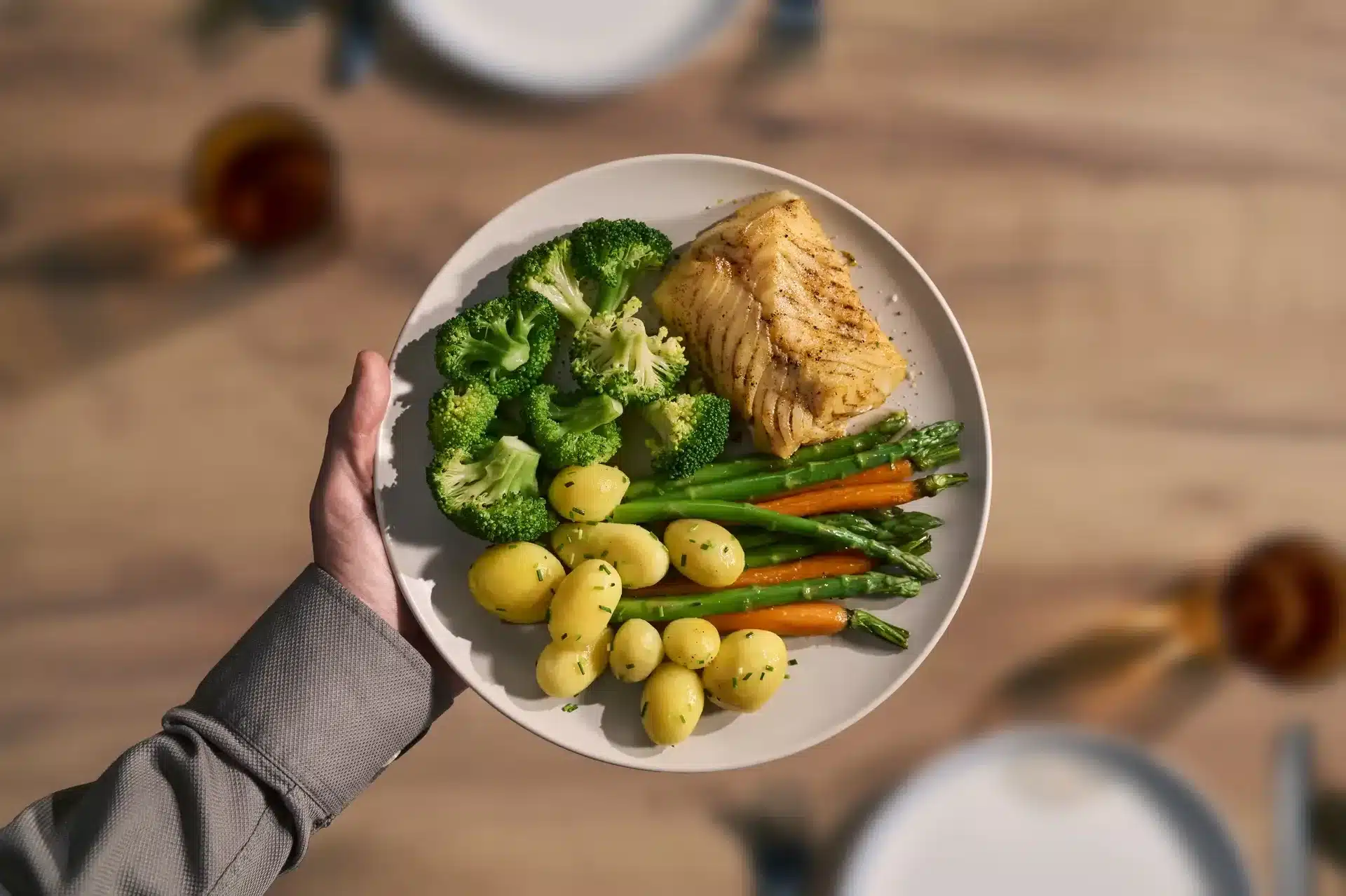 Teller mit gegarten Kartoffeln, Brokkoli, grünem Spargel, Karotten und einem Stück Fischfilet, in der Hand gehalten, von oben fotografiert.
