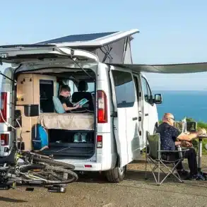 Antilope Van Flex 5 Campingbus mit offenem Heck, aufgeklapptem Aufstelldach, Fahrrädern und Personen beim Outdoor-Frühstück am Meer.