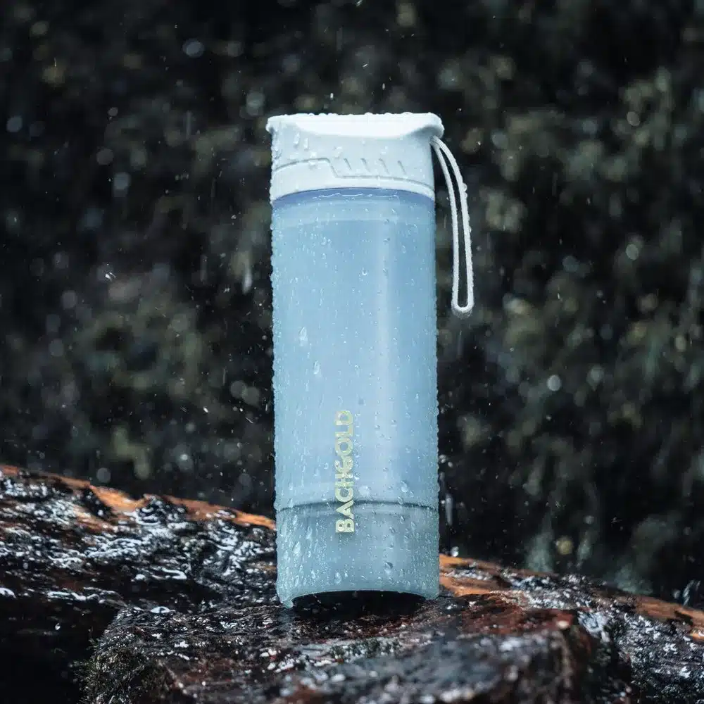Der Bachgold® Selfpress Wasserfilter als Outdoor-Trinkflasche, stehend auf einem nassen Stein mit Wassertropfen.