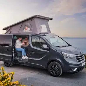 Grauer Renault Trafic Campervan mit Aufstelldach, geparkt an einer Küste; vier Personen entspannen im und am Fahrzeug