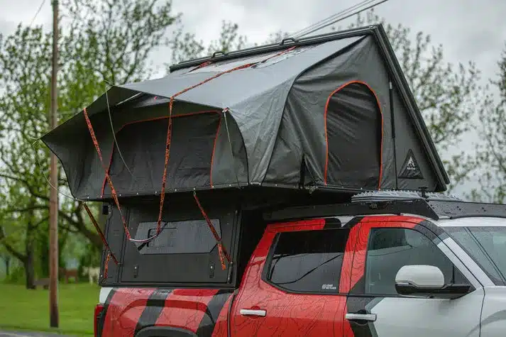 Alu-Cab ModCAP Family Camper mit seitlich geöffnetem Dachzelt auf Pick-up-Ladefläche bei regnerischem Wetter.
