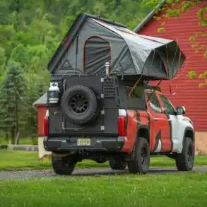 Pick-up mit Alu-Cab ModCAP Family Camper-Aufbau und geöffnetem Aufstelldach auf einer grünen Wiese vor roter Scheune.