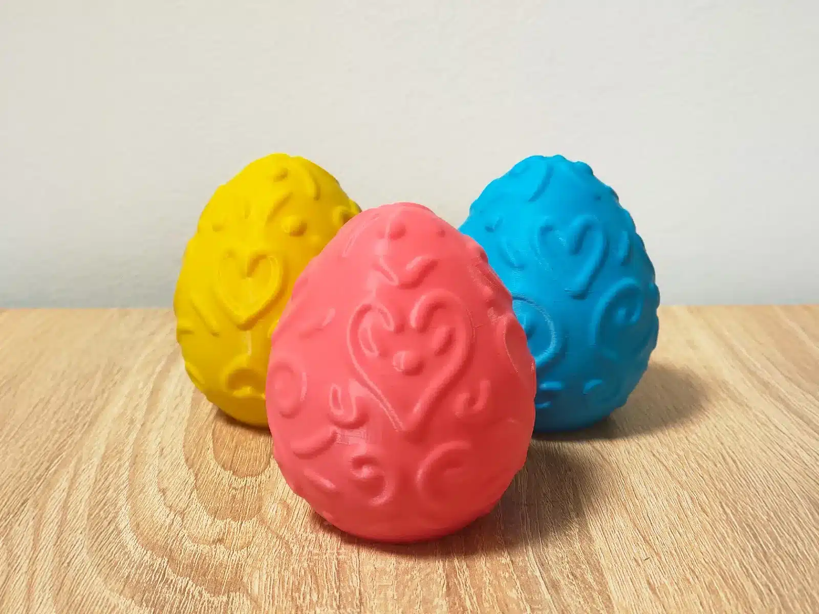 Drei bunte 3D-gedruckte Eier mit Herzmustern auf hellem Holztisch