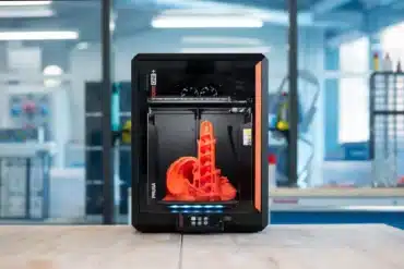 Frontalansicht des Prusa CORE One+ 3D-Druckers mit laufendem Druck eines orangefarbenen Teils in einer Werkstattumgebung.