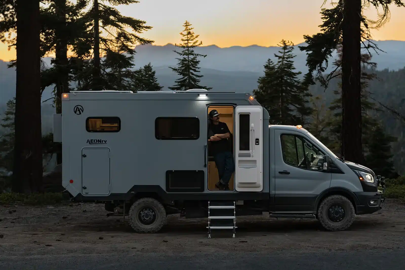 Seitenansicht eines AEONrv Offroad-Campers mit geöffneter Tür und Mensch im Eingang, im Wald bei Sonnenuntergang.
