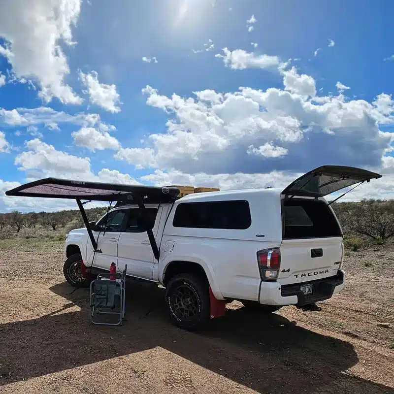 Weiße Tacoma-Pickup mit aufgebauter AirHaven 40 Markise von DAYLODGE und geöffneter Heckklappe auf Campingplatz.