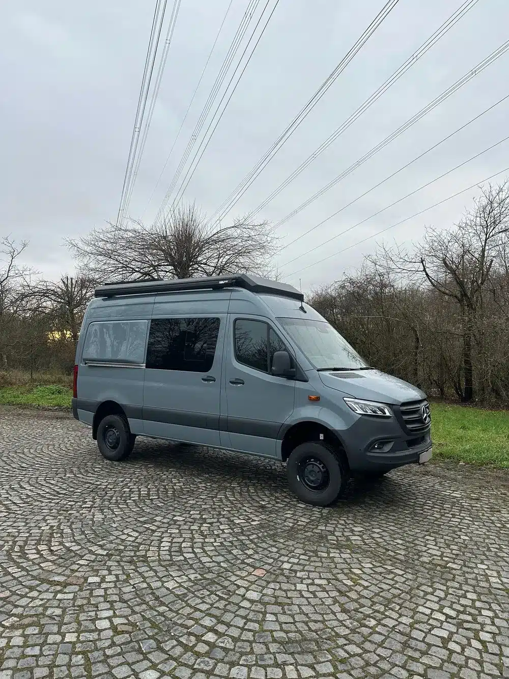 Grauer Mercedes Sprinter mit geschlossenem Campany-Aufstelldach von Reisewagenworks auf einem gepflasterten Platz.