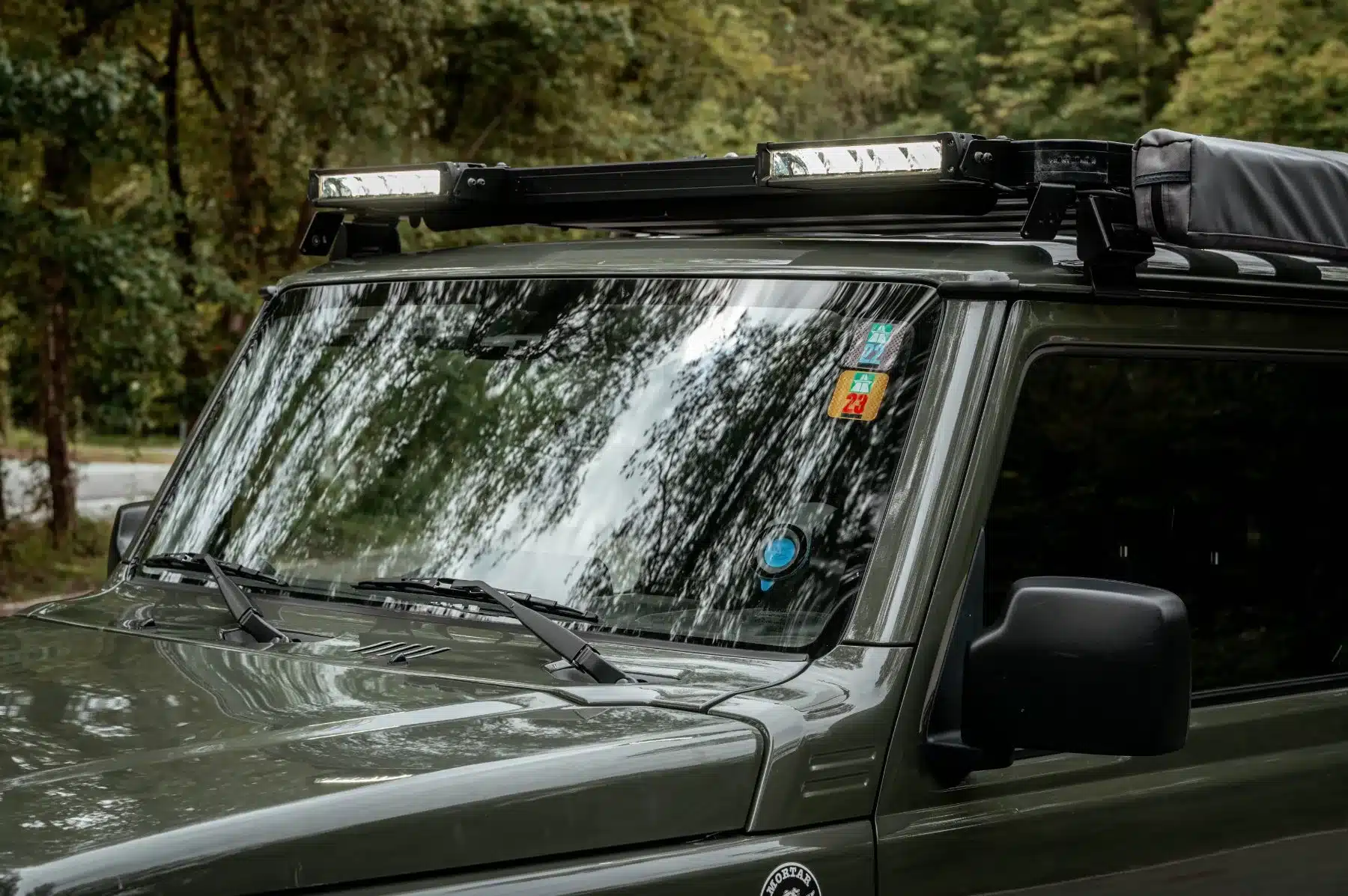 Frontansicht eines Suzuki Jimny mit Dachträger, LED-Zusatzscheinwerfern und reflektierender Frontscheibe im Waldgebiet.