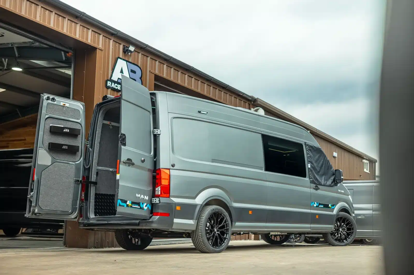 Grauer AB Race Cruisers Campervan John McGuinness Edition mit geöffneten Hecktüren, vor Werkstatthalle geparkt.