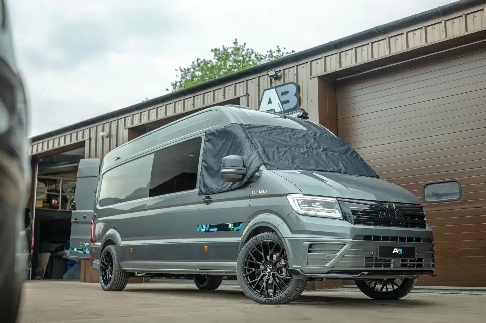 AB Race Cruisers John McGuinness Edition Sondermodell Kastenwagen in Grau vor Produktionshalle mit schwarzen Alufelgen und Sichtschutz an Frontscheibe