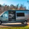 Seitenansicht eines MAN TGE 180 Campervans mit geöffneter Schiebetür, sichtbarer Küchenzeile und Outdoor-Umgebung.