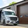 Mercedes Sprinter 319 V6 von AB Race Cruisers mit langem Radstand, geöffneter Schiebetür, vor einer Werkstatthalle.