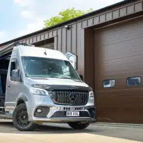 Mercedes Sprinter 319 V6 von AB Race Cruisers mit langem Radstand, geöffneter Schiebetür, vor einer Werkstatthalle.