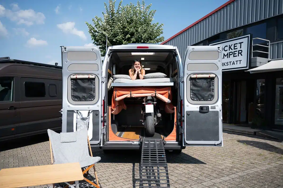 Heckansicht eines Rocket Moto Campervans mit aufgeklapptem Heck, Motorrad und hochgefahrenem Hubbett, Frau liegt im Heckbereich.