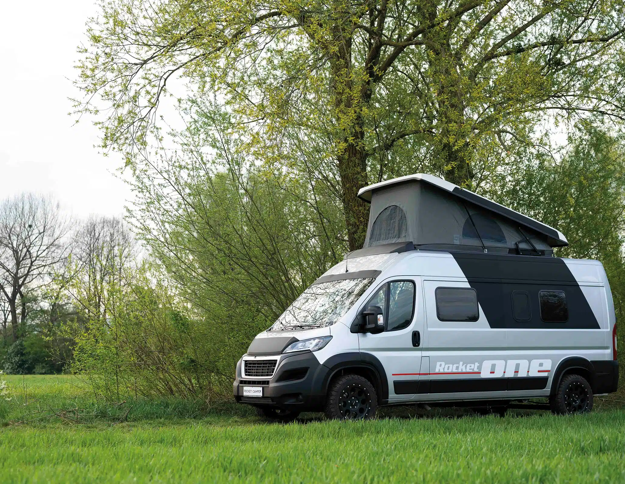Der ROCKET ONE M Campervan mit ausgefahrenem Aufstelldach steht auf einer Wiese vor Bäumen.