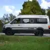 Seitenansicht des Rocket TGE Kastenwagens von Rocket Camper, geparkt auf einer Landstraße vor grüner Natur.