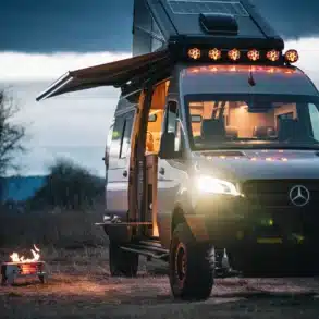 Skyloft Van von Redtail Overland mit festem Dachschlafzimmer, aufgeklappter Markise und Lagerfeuer im Vordergrund bei Dämmerung.