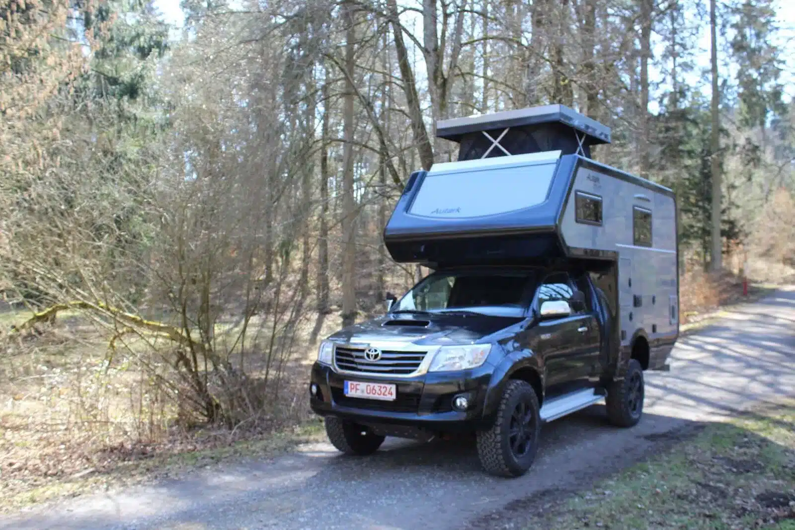 Schwarzer Toyota Hilux mit grauer Autark-Wohnkabine und geöffnetem Hubdach auf einem Waldweg
